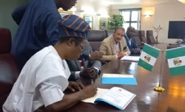 Marvelous Mike Press Limited chief executive officer, Michael Akinola signing agreement watched by Tourism Minister, Charles Max Fernandez in brown suit.